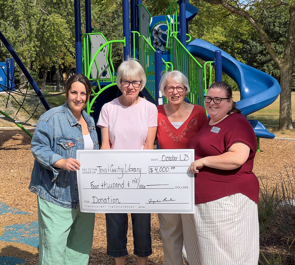 Four members of the Friends of the Library pose in front of a playground holding a giant check